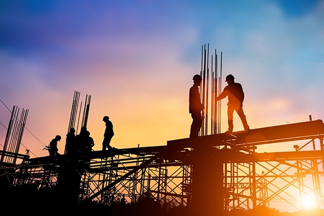 Silhouetted construction workers stand tall on scaffolding, framed by a vibrant sunset sky